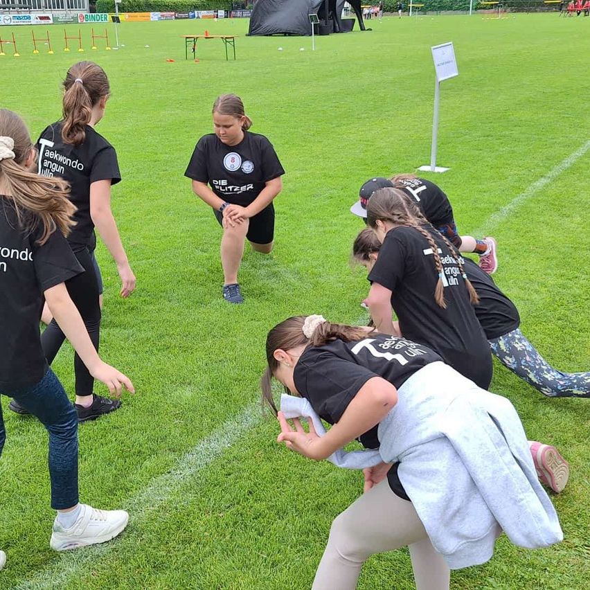Eine Gruppe junger Mädchen in Sportbekleidung trainiert auf einem Grasfeld. Eine Mädchen beugt sich über, um ihren Schuh zu schnüren, während andere zusehen. Einige Hürden sind auf dem Feld aufgestellt.