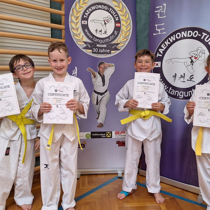Vier Jungen in weißen Taekwondo-Uniformen mit gelben Gürteln stehen zusammen und halten Urkunden in der Hand. Sie lächeln für ein Foto vor einem lila Hintergrund mit Kampfkunstgrafiken.