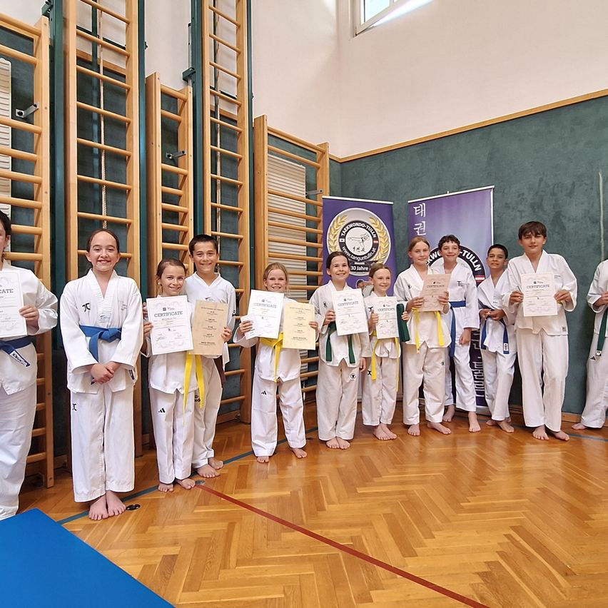 Eine Gruppe von Kindern, in Karate-Uniformen gekleidet, steht in einem Fitnessstudio und hält Urkunden in den Händen. Sie lächeln für ein Foto. Der Raum hat Holzböden und Kletterleitern an den Wänden.