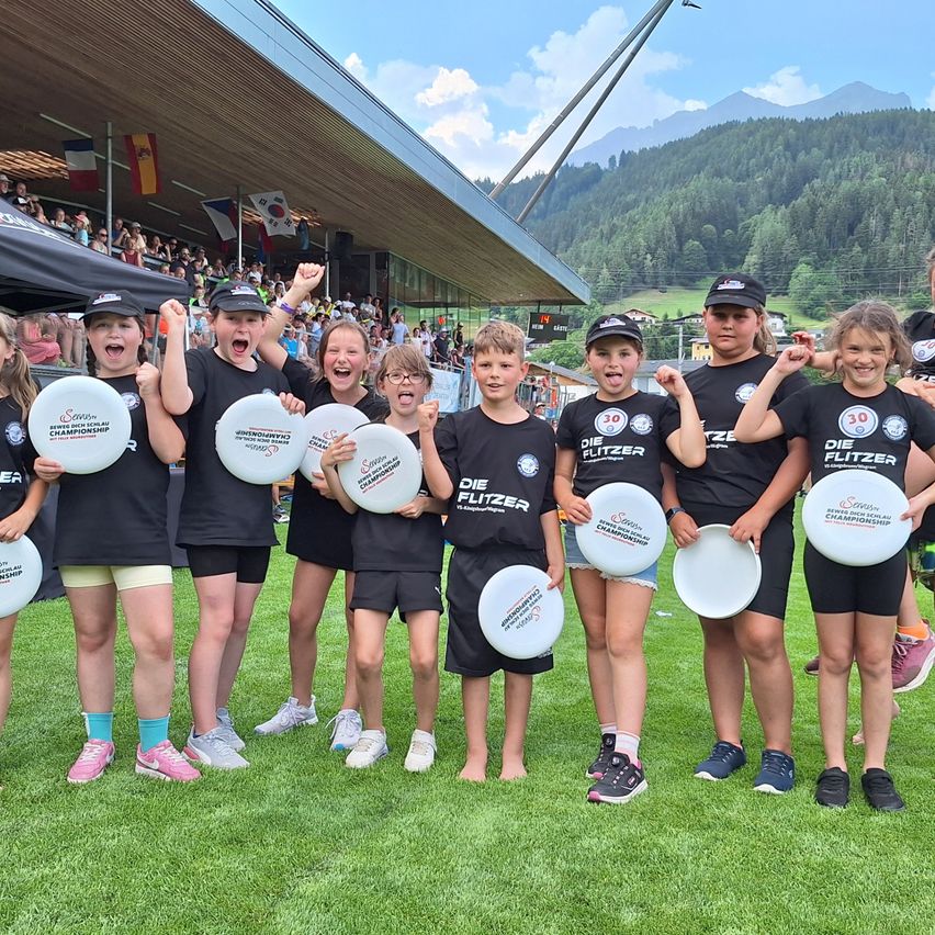 Eine Gruppe von Kindern in gleichen schwarzen T-Shirts und Shorts steht auf einem grünen Feld und hält weiße Frisbees in der Hand, mit Bergen und einem Stadion im Hintergrund.
