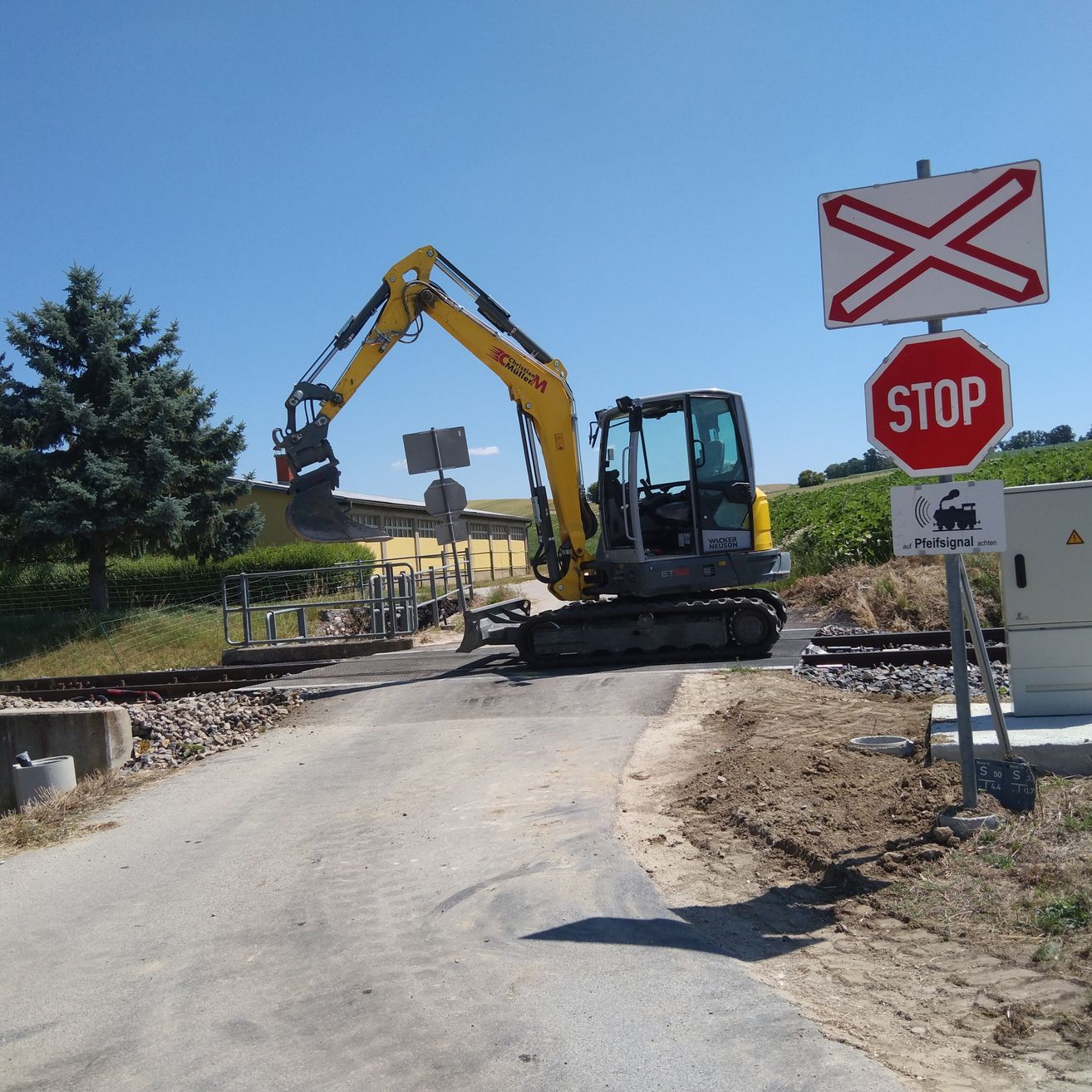 Ein gelber Bagger steht in der Nähe eines Stopp-Schildes und eines Bahnübergangs. Der Bagger befindet sich auf einer Straße mit Kies und einem Baum im Hintergrund.