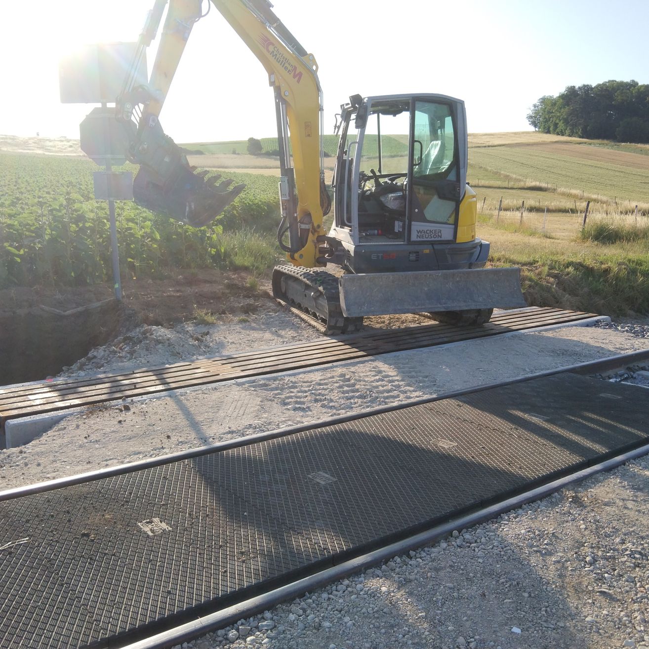 Ein gelber Bagger steht neben einer Metallgitterplatte auf einem Schotterweg, mit einem grünen Feld und Bäumen im Hintergrund.