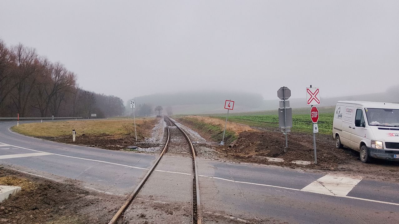 Eine Straße überquert unter einem nebligen Himmel eine Eisenbahnstrecke. Die Strecke hat zwei Schilder, eines mit der Nummer 24 und das andere mit der Nummer 4.