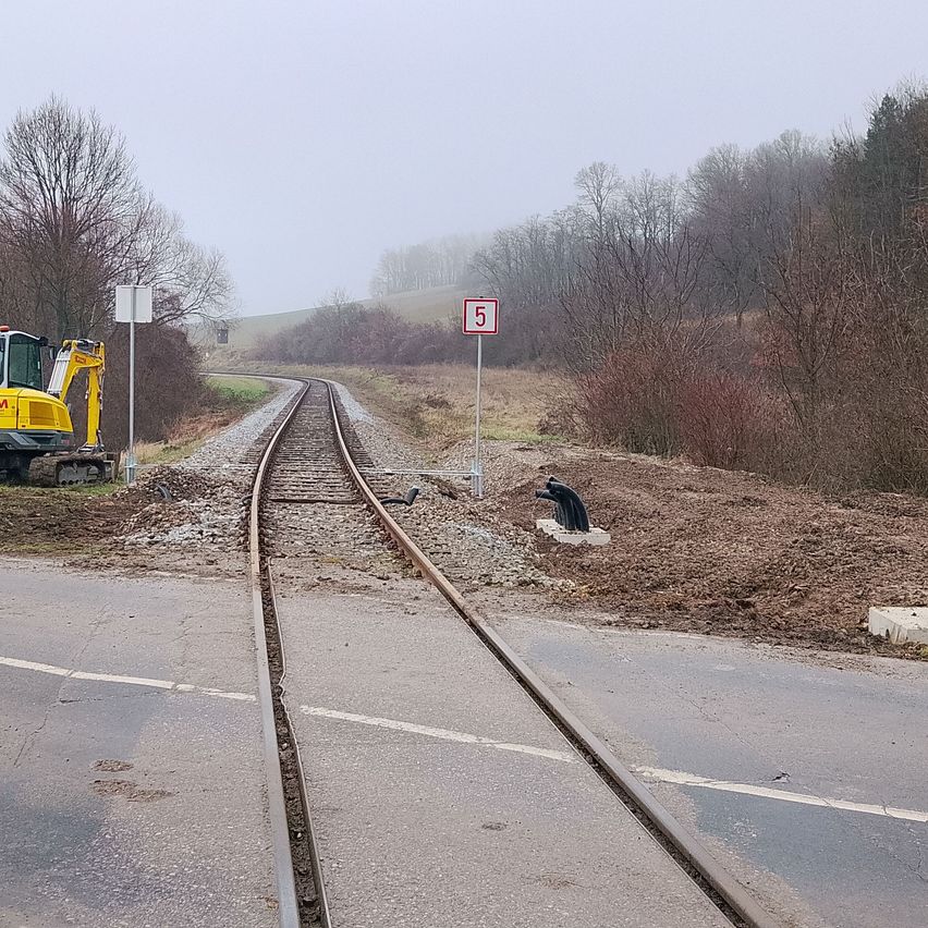 Eine Eisenbahnstrecke mit einem Nummer-5-Schild. Ein gelber Bagger steht links. Bäume und Büsche umgeben das Gebiet. Der Boden ist nass.