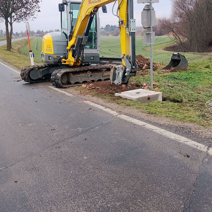 Ein gelber und grauer Bagger steht am Straßenrand, mit einem vollen Eimer Erde daneben. Ein Verkehrsschild befindet sich auf der Rasenfläche. Die Straße ist leer.