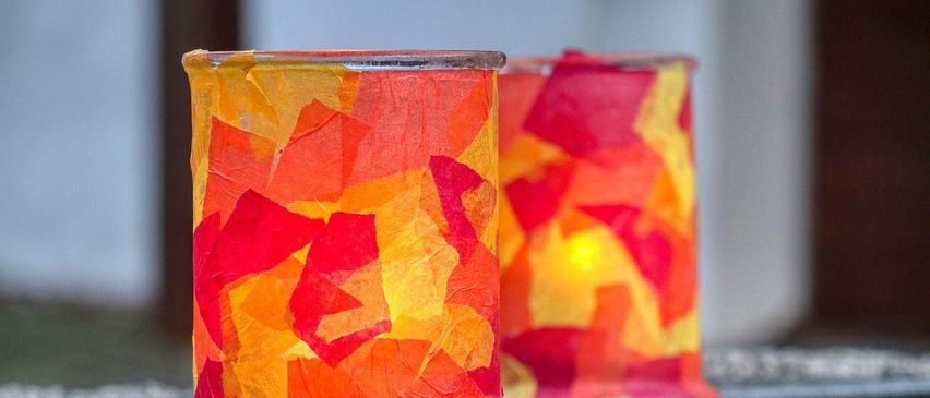 Zwei handgefertigte dekorative Kerzenhalter, verziert mit leuchtenden roten und gelben Papierausschnitten, stehen auf einer Metalloberfläche. Der Hintergrund ist unscharf.