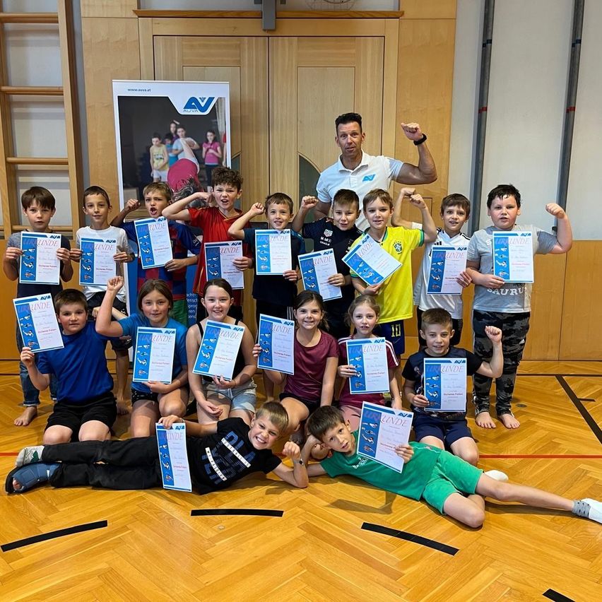 Eine Gruppe von Kindern mit Urkunden posiert mit einem Trainer in einer Turnhalle.
