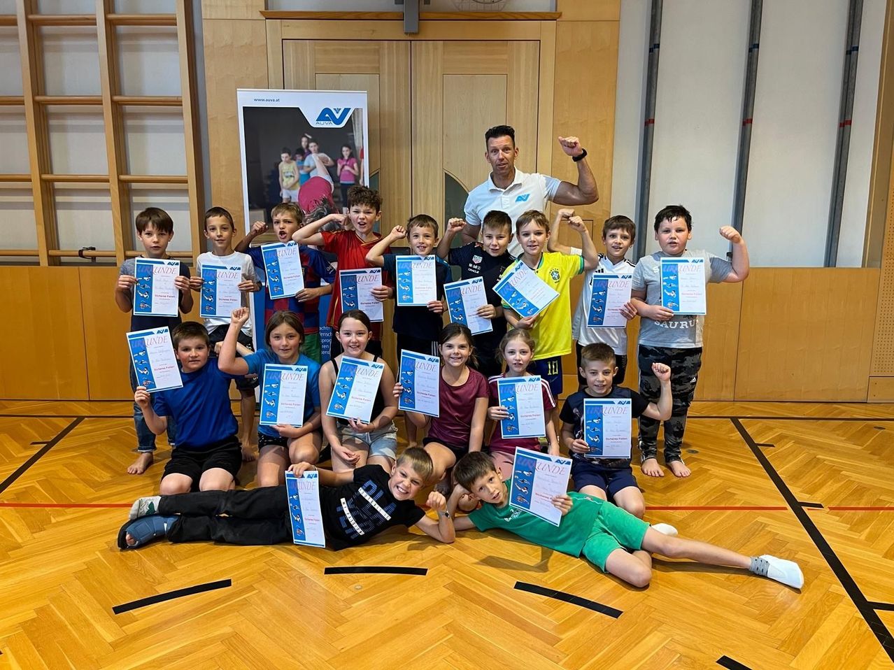 Eine Gruppe von Kindern mit Urkunden posiert mit einem Trainer in einer Turnhalle.