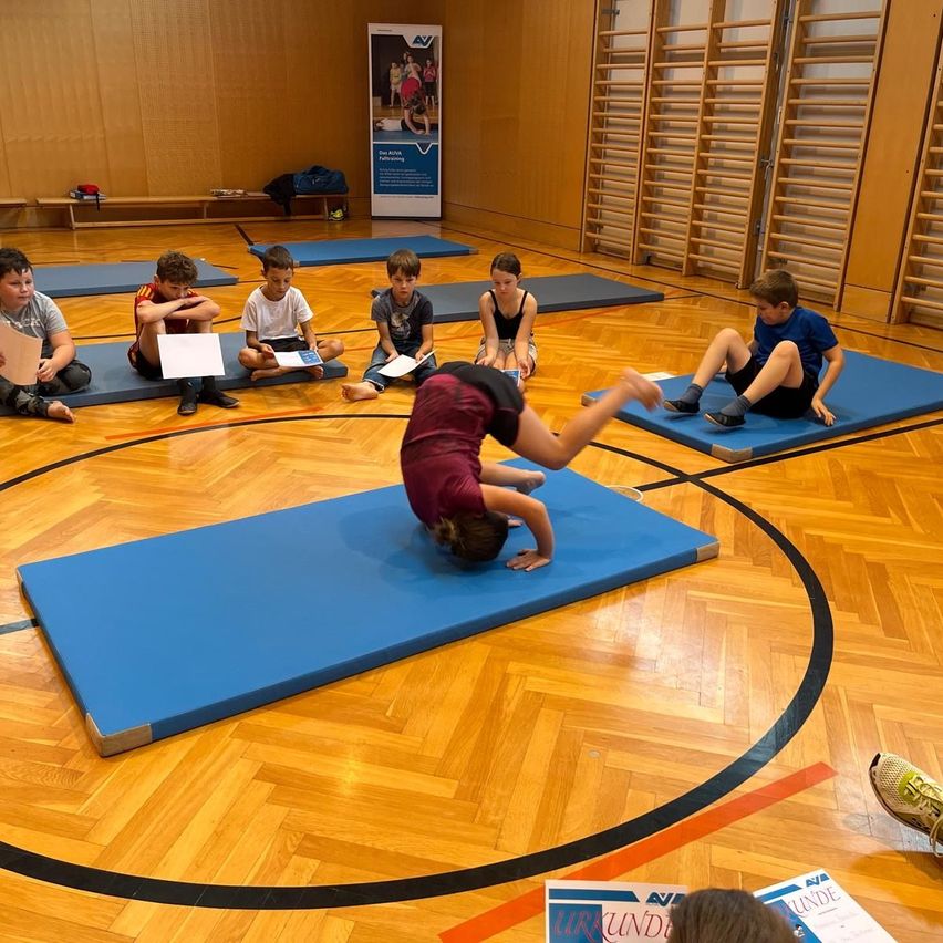 Eine Gruppe von Kindern macht Yoga auf blauen Matten in einer Turnhalle. Ein Kind macht einen Handstand, während die anderen sitzen und zuschauen.