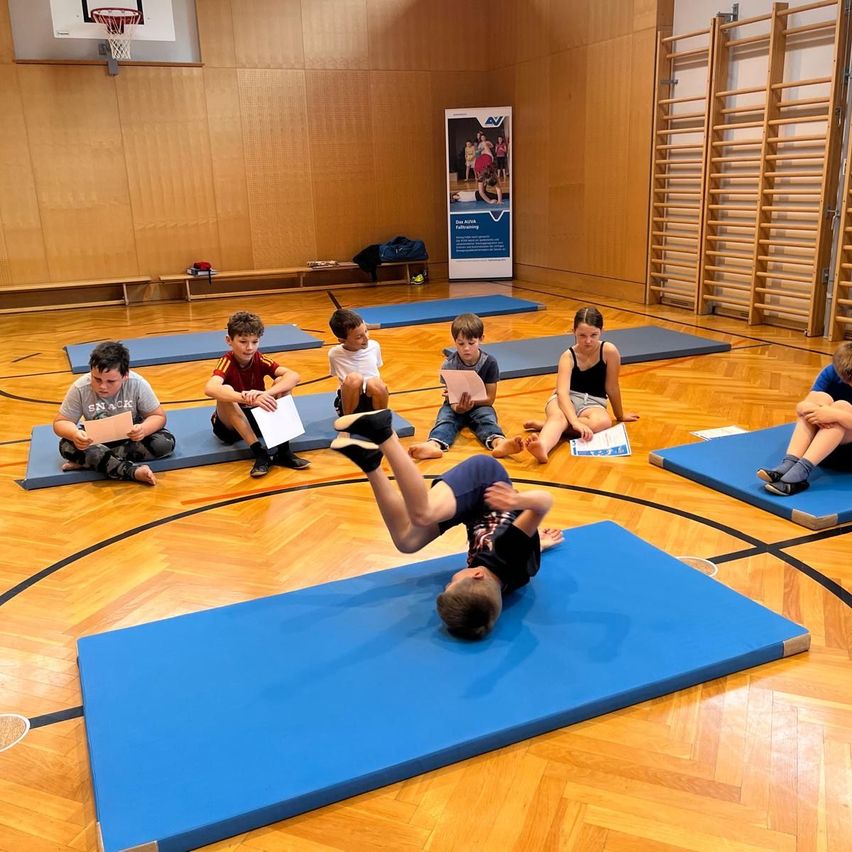 Kinder üben Gymnastik auf blauen Matten in einer Turnhalle. Ein Kind macht einen Handstand. Andere beobachten oder lesen Papiere. Holzwände und Geräte sind im Hintergrund.