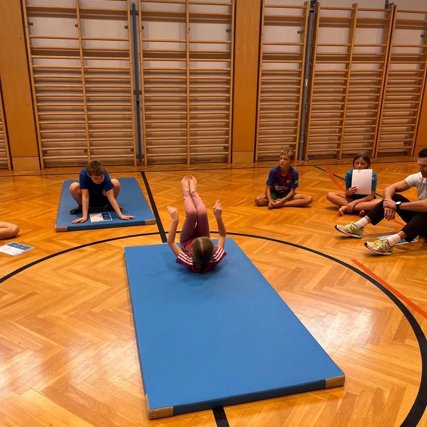 Eine Gruppe von Kindern übt Gymnastik auf blauen Matten in einer Turnhalle, während andere zuschauen. Sie tragen Turnschuhe.