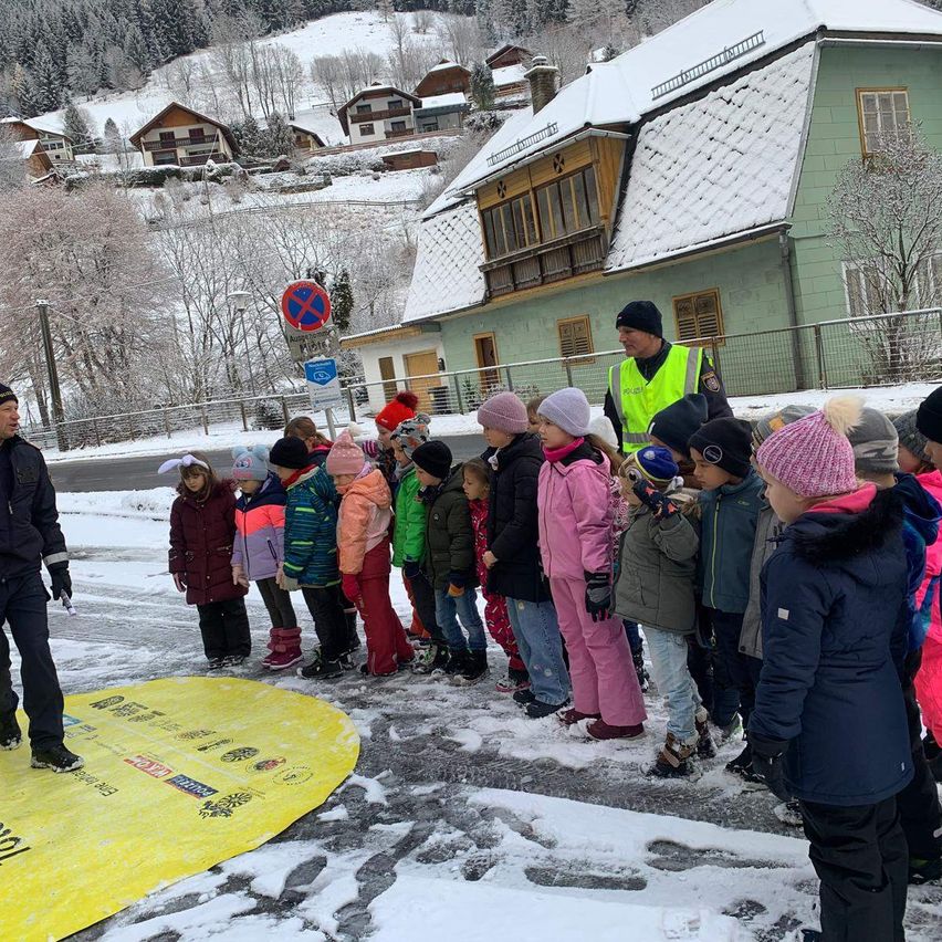 Eine Gruppe von Kindern in Winterkleidung steht in einem verschneiten Bereich, wobei ein Mann in einer Warnweste sie anführt. Ein gelbes Schild liegt vor ihnen auf dem Boden.
