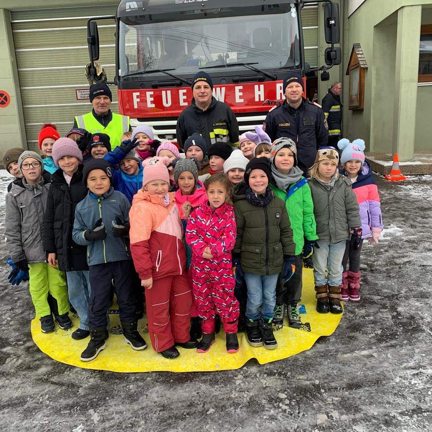Eine Gruppe von Kindern und Erwachsenen steht auf einem gelben Teppich vor einem Feuerwehrauto, mit einem Feuerwehrmann in Uniform in der Mitte.