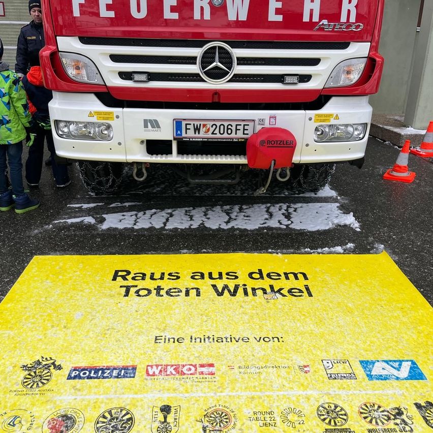 Ein roter Feuerwehrwagen mit der Aufschrift 'Feuerwehr' ist auf einer verschneiten Straße geparkt. Darunter steht auf einem gelben Teppich 'Raus aus dem Toten Winkel'. Einige Leute stehen um den Wagen herum. Orangefarbene Verkehrskegel sind auf der Straße aufgestellt.