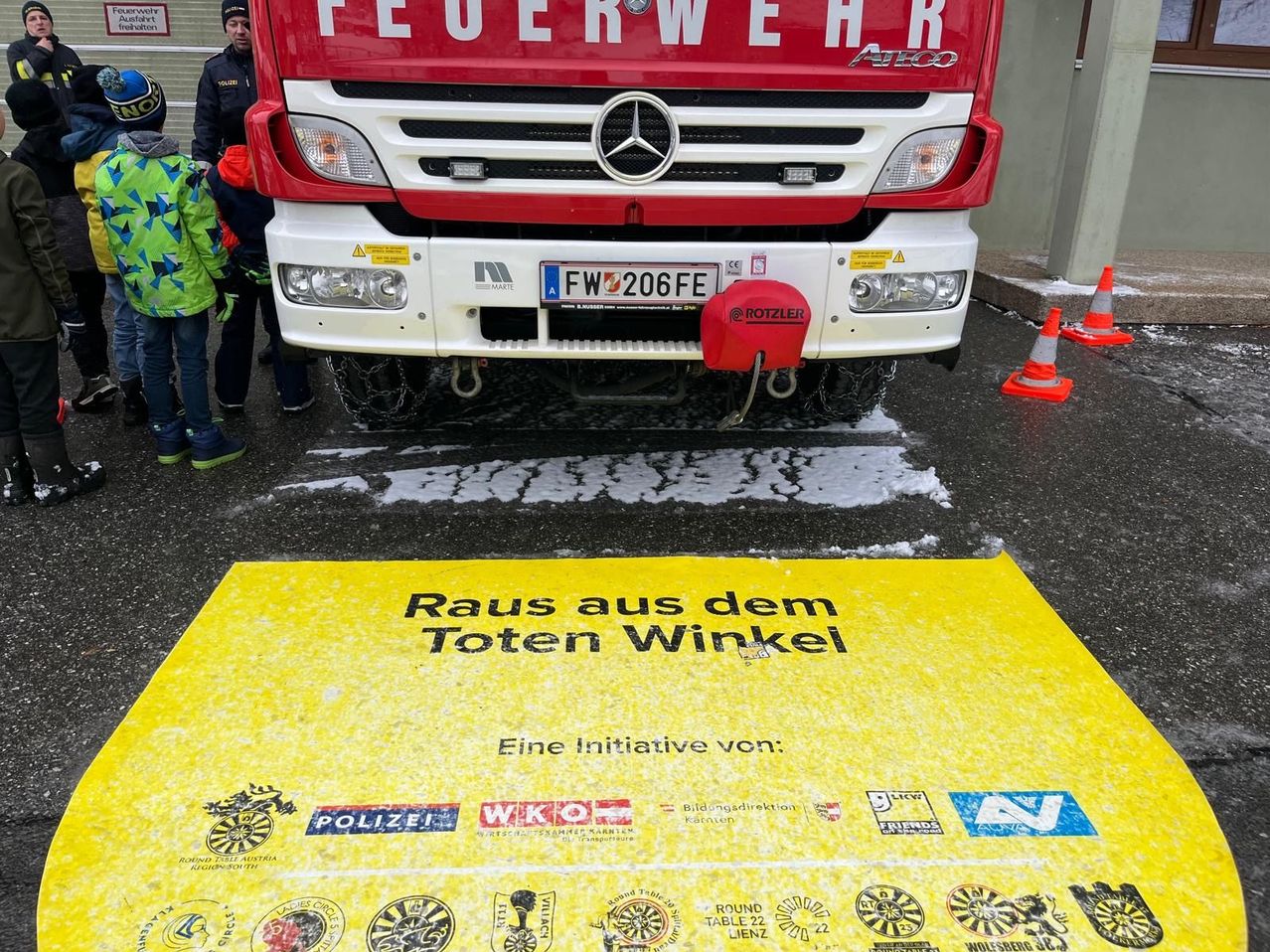 Ein roter Feuerwehrwagen mit der Aufschrift 'Feuerwehr' ist auf einer verschneiten Straße geparkt. Darunter steht auf einem gelben Teppich 'Raus aus dem Toten Winkel'. Einige Leute stehen um den Wagen herum. Orangefarbene Verkehrskegel sind auf der Straße aufgestellt.