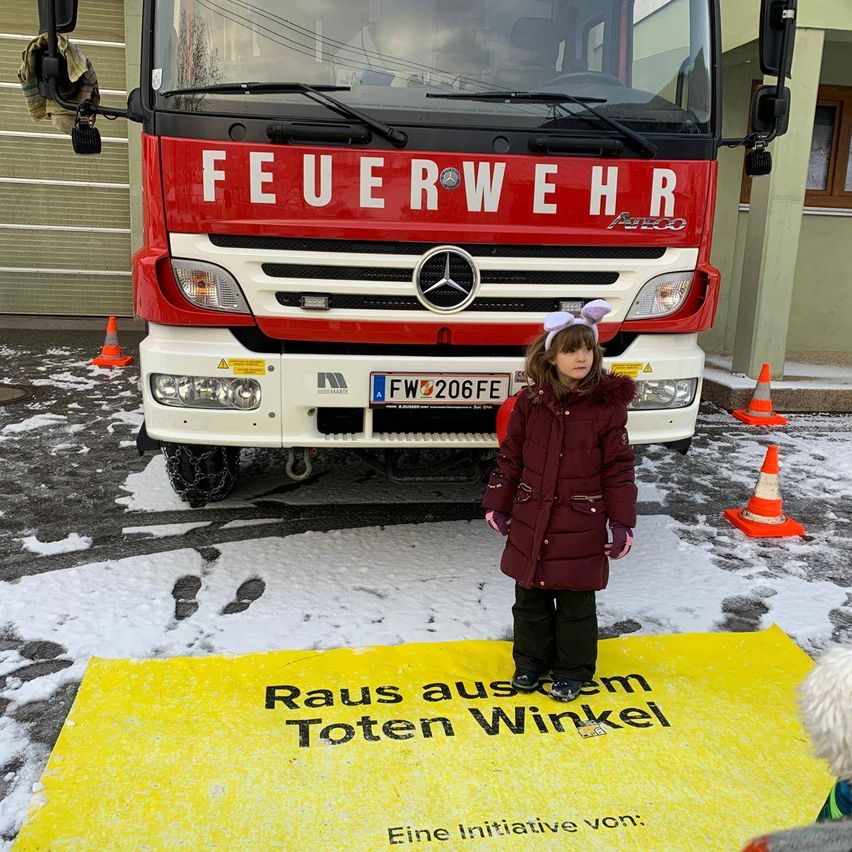 Ein Mädchen in einem Mantel steht vor einem Feuerwehrauto im Schnee. Auf dem Auto steht 'FEUERWEHR' und das Mädchen trägt Hasenohren.
