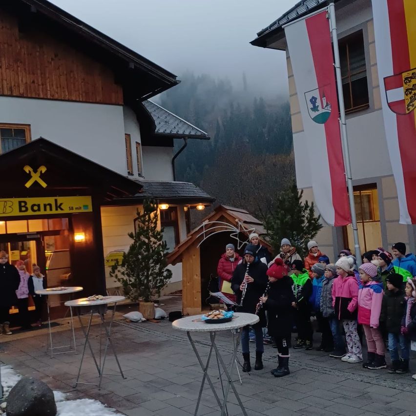 Eine Gruppe von Menschen, darunter Kinder, versammelt sich vor einem Gebäude mit Fahnen, einer von ihnen spielt ein Musikinstrument. Dahinter steht eine kleine Hütte und eine Bank mit einem Tisch davor.
