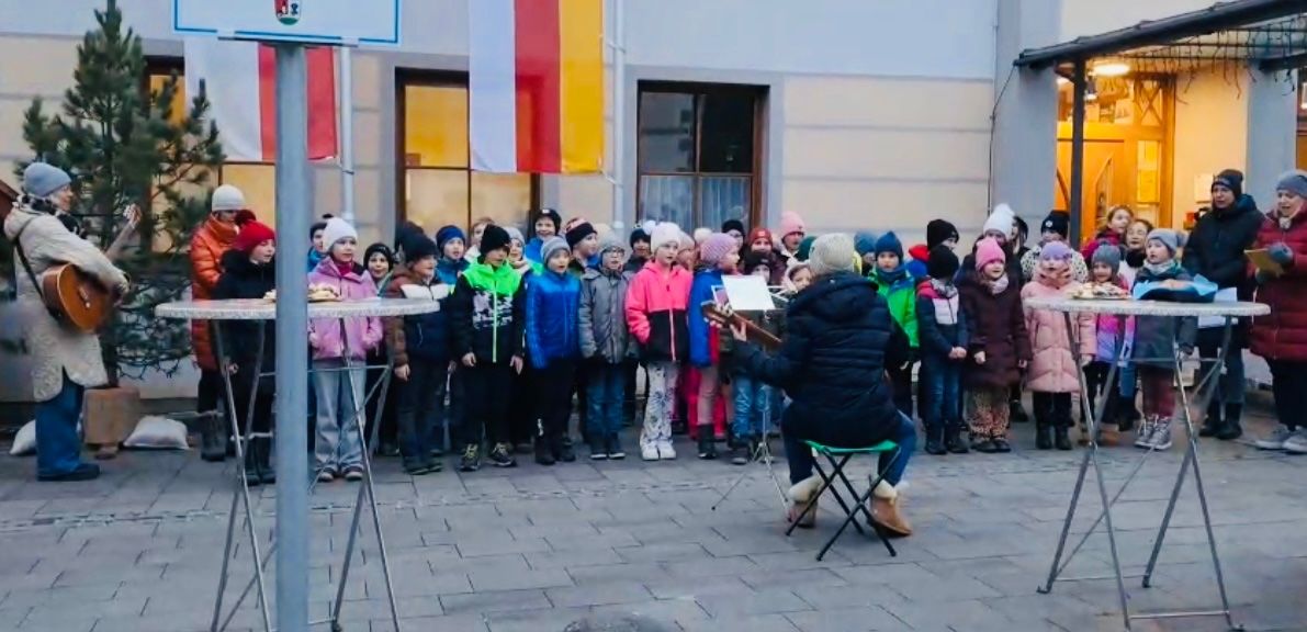 Eine Gruppe von Kindern in Winterkleidung steht in einer Reihe und singt. Eine Person in einem schwarzen Mantel sitzt auf einem grünen Stuhl und spielt Gitarre. Ein Gebäude mit Fahnen und Fenstern steht dahinter.