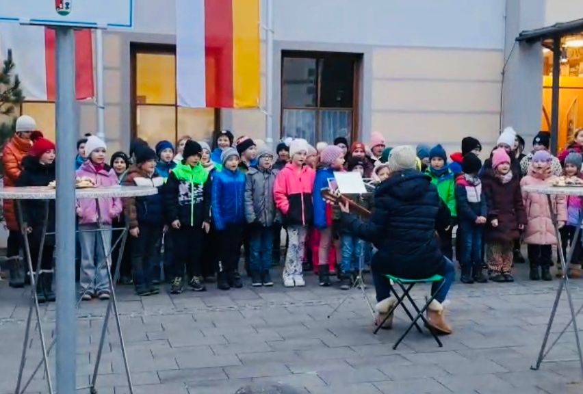 Eine Gruppe von Kindern in Winterkleidung steht in einer Reihe und singt. Eine Person in einem schwarzen Mantel sitzt auf einem grünen Stuhl und spielt Gitarre. Ein Gebäude mit Fahnen und Fenstern steht dahinter.