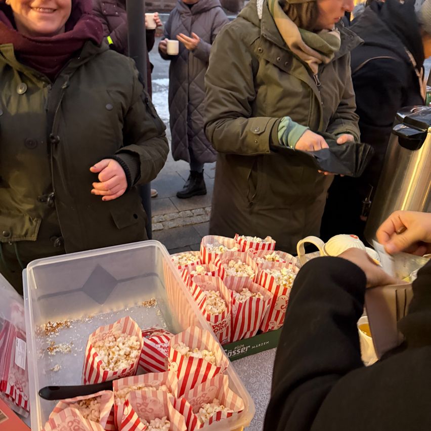 Eine Gruppe von Menschen steht draußen, wobei eine Person Popcorn aus einem Behälter serviert. Zwei Frauen tragen Mäntel und Schals, und einige halten Tassen. Der Tisch ist mit Popcorn in rot-weiß gestreiften Tüten bedeckt.