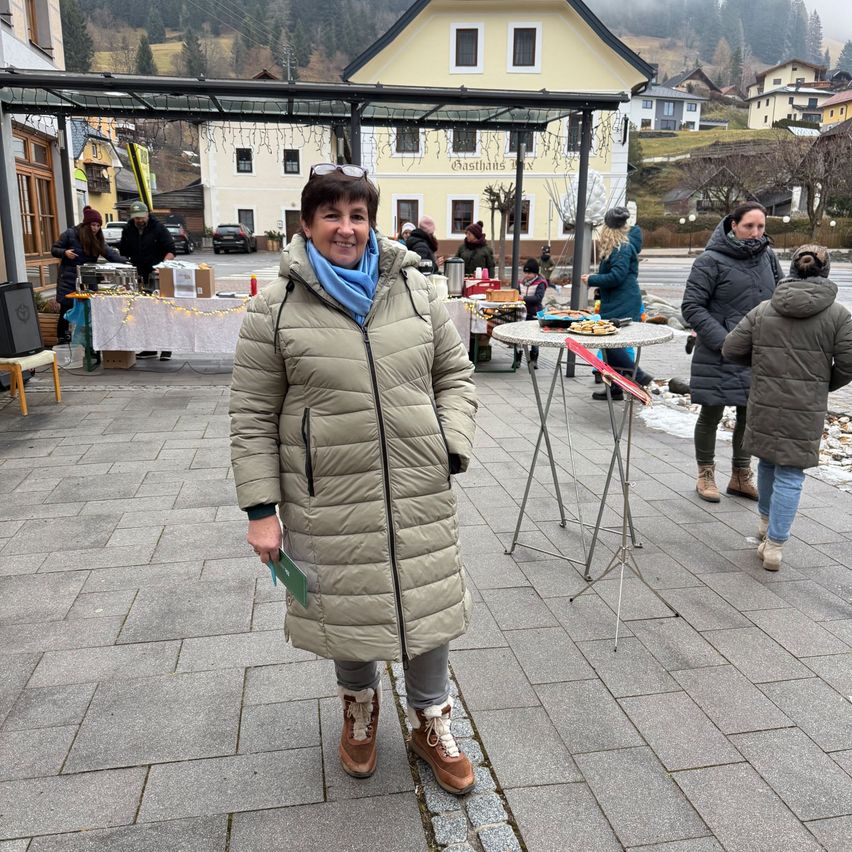 Eine Frau steht auf einer gefliesten Straße, lächelt und trägt einen beigen Mantel und einen blauen Schal. Hinter ihr stehen Tische mit Essen, und andere Leute gehen vorbei. Im Hintergrund befinden sich Gebäude, Bäume und ein Berg.