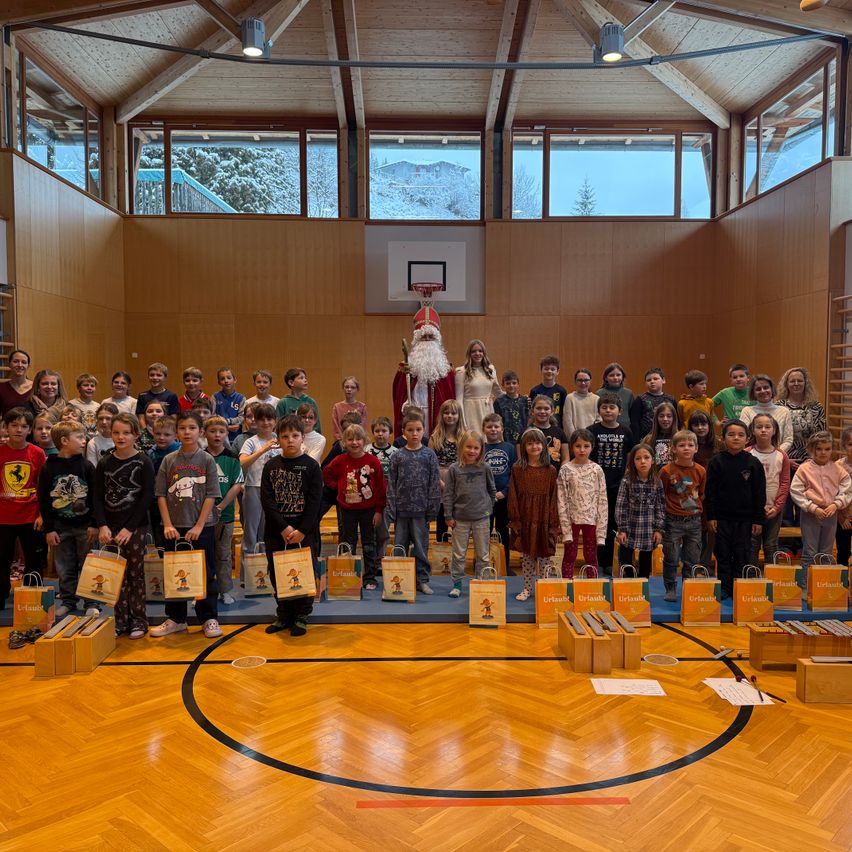 Eine Gruppe von Kindern steht in einer Turnhalle, hält Geschenktüten und hat den Weihnachtsmann in der Mitte. Die Turnhalle hat einen Holzboden, Holzwände und große Fenster. Im Hintergrund ist ein Basketballkorb zu sehen.
