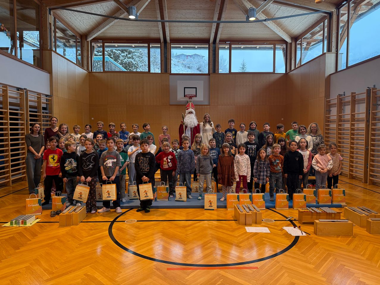 Eine Gruppe von Kindern steht in einer Turnhalle, hält Geschenktüten und hat den Weihnachtsmann in der Mitte. Die Turnhalle hat einen Holzboden, Holzwände und große Fenster. Im Hintergrund ist ein Basketballkorb zu sehen.
