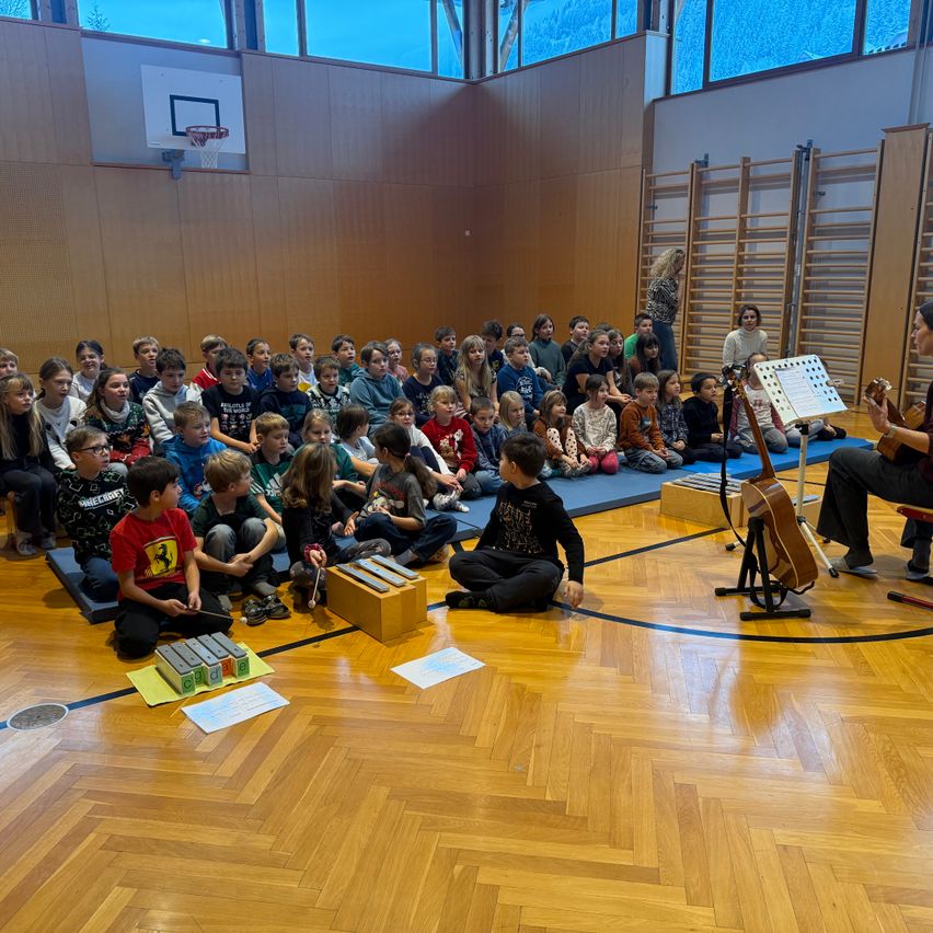 Kinder sitzen im Halbkreis auf dem Hallenboden und schauen einem Menschen zu, der eine Gitarre spielt und einen Notenständer mit Noten vor sich hat. Vor den Kindern liegen Xylophon und Papiere. Im Hintergrund ist ein Basketballkorb zu sehen.