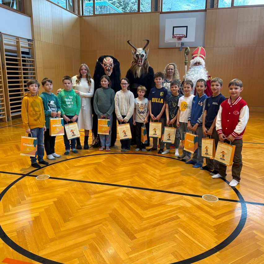 Eine Gruppe von Kindern und zwei Erwachsenen steht in einer Turnhalle. Die Kinder halten Geschenktüten, während zwei Erwachsene als Krampus und Weihnachtsmann verkleidet sind. Dahinter ist ein Basketballkorb an der Wand befestigt.