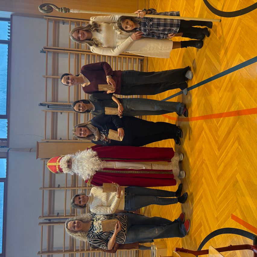 Eine Gruppe Erwachsener in festlichen Kostümen, möglicherweise zur Feier eines Feiertags, stehen zusammen in einer Turnhalle. Einer, als Weihnachtsmann verkleidet, hält ein Buch. Sie lächeln und posieren für ein Foto.