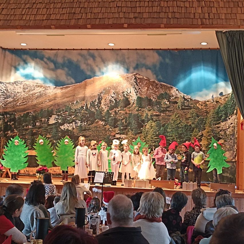 Eine Gruppe von Kindern, die als Weihnachtsbäume verkleidet sind, treten auf einer Bühne mit einem Berghintergrund auf. Das Publikum sitzt vor der Bühne.