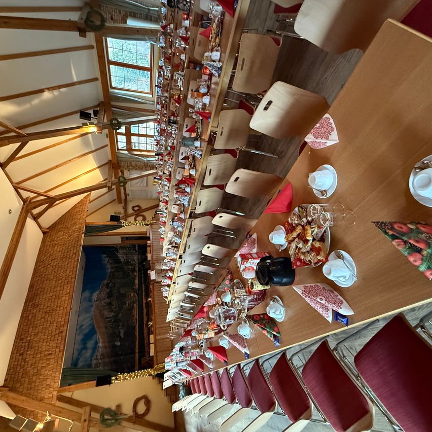 Ein Bankettsaal mit hölzernen Deckenbalken, mehreren langen Tischen, die mit roten Servietten, Tassen, Untertassen und verschiedenen Speisen gedeckt sind. Die Tische sind mit Stühlen angeordnet, und es gibt einen großen dekorativen Hintergrund mit einer Bergszene.