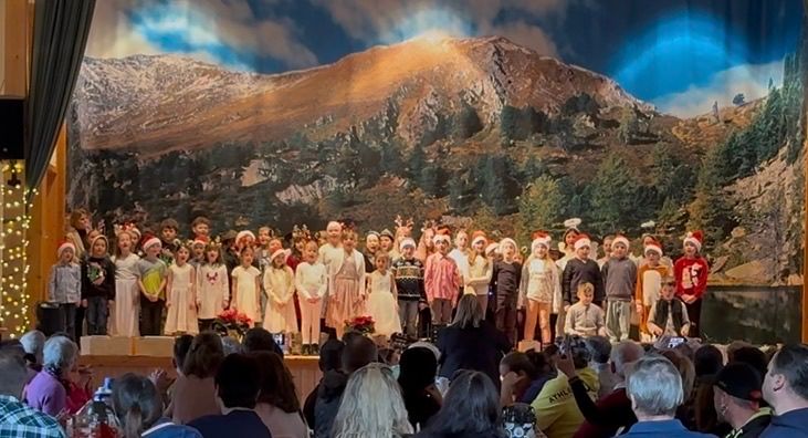 Ein Chor von Kindern in festlicher Kleidung tritt auf der Bühne auf, mit einem Berg im Hintergrund und einer Menge erwachsener Zuschauer.