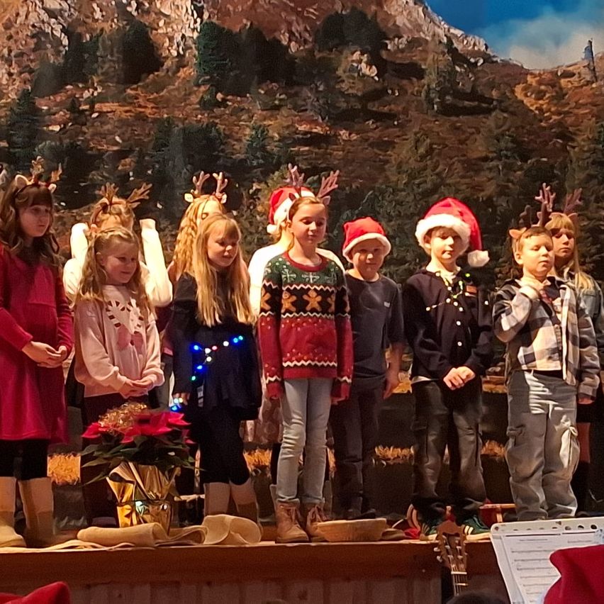 Eine Gruppe von Kindern in Weihnachtskleidung steht auf der Bühne. Einige tragen Rentier-Kopfbedeckungen und einige haben Weihnachtsmützen. Vor ihnen steht ein Blumenarrangement und eine Person hält eine Gitarre auf der Bühne.