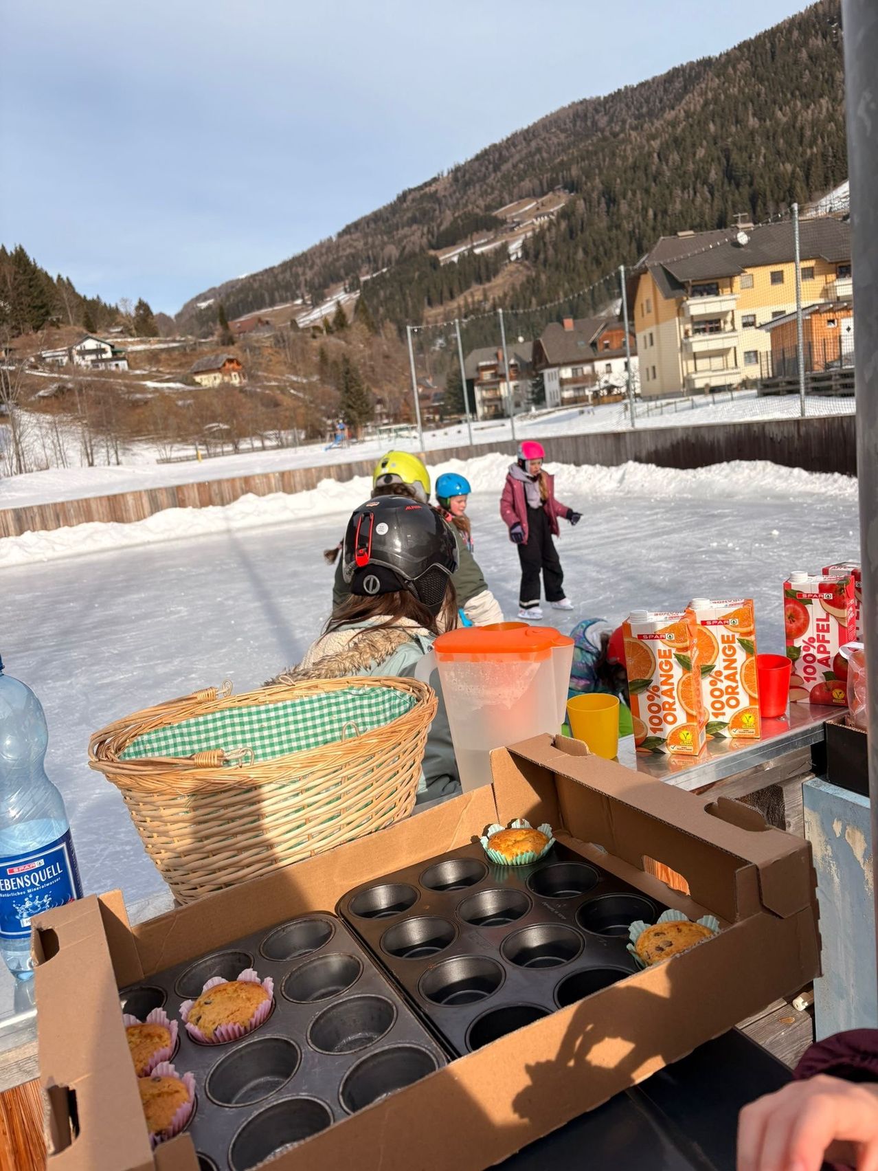 Eine verschneite Landschaft mit Bergen im Hintergrund, Kinder sitzen auf dem Eisplatz und tragen Helme, und rechts befindet sich ein Tisch mit Essen und Getränken.