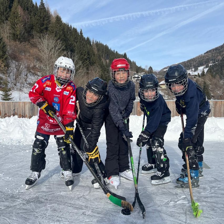 Fünf junge Eishockeyspieler posieren für ein Foto auf einer Eisbahn. Sie tragen alle Helme und halten Eishockeyschläger. Der Linke hat ein rotes Trikot.