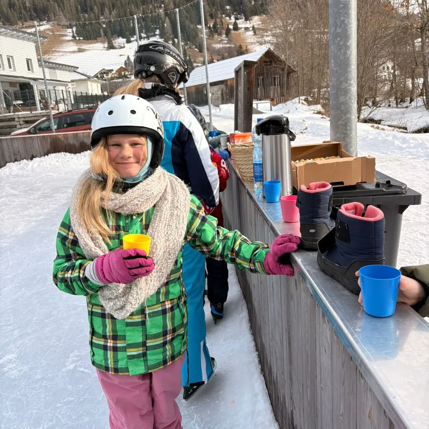 Ein junges Mädchen in Winterkleidung steht an einer Theke und hält einen gelben Becher in der Hand, mit anderen Personen im Hintergrund.