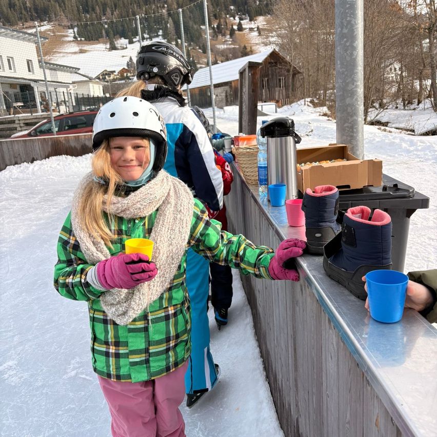 Ein junges Mädchen in Winterkleidung steht an einer Theke und hält einen gelben Becher in der Hand, mit anderen Personen im Hintergrund.