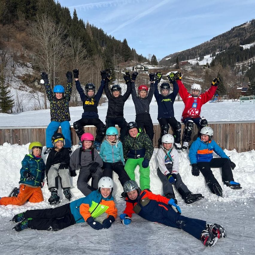 Eine Gruppe von Kindern in Winterkleidung posiert für ein Foto auf einer Eisbahn, mit verschneiten Bergen im Hintergrund.
