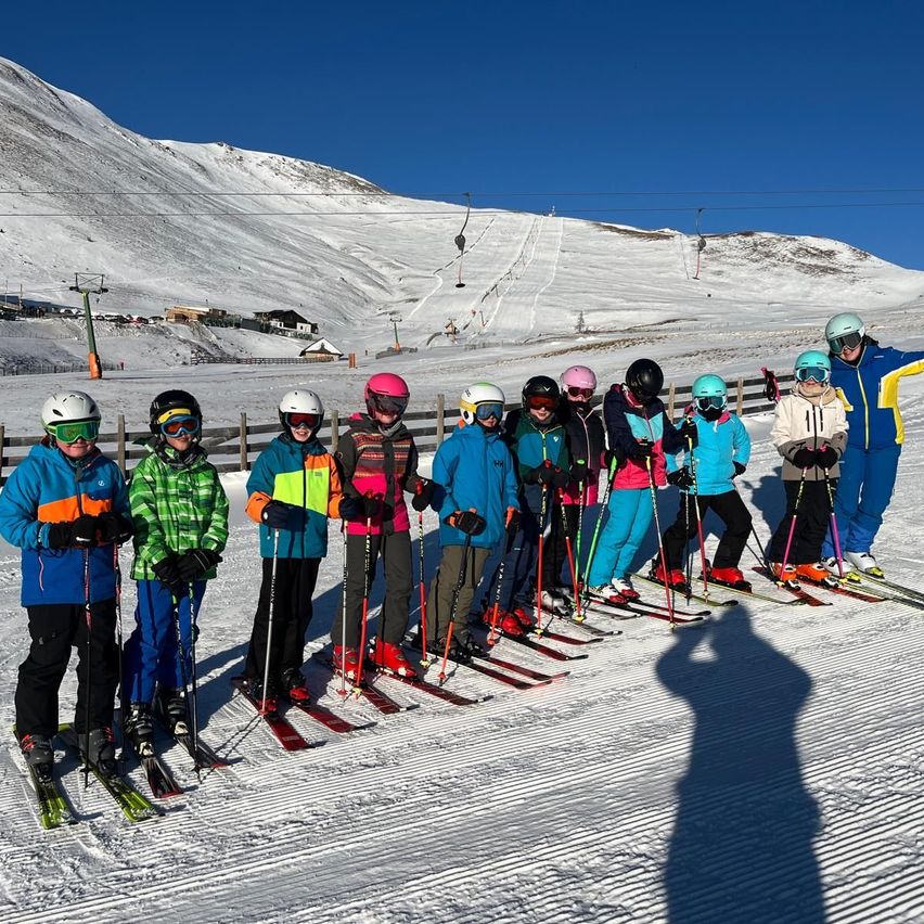 Eine Gruppe von Skifahrern posiert für ein Foto auf einem verschneiten Berg, mit klarem blauem Himmel oben und Skiliften im Hintergrund.