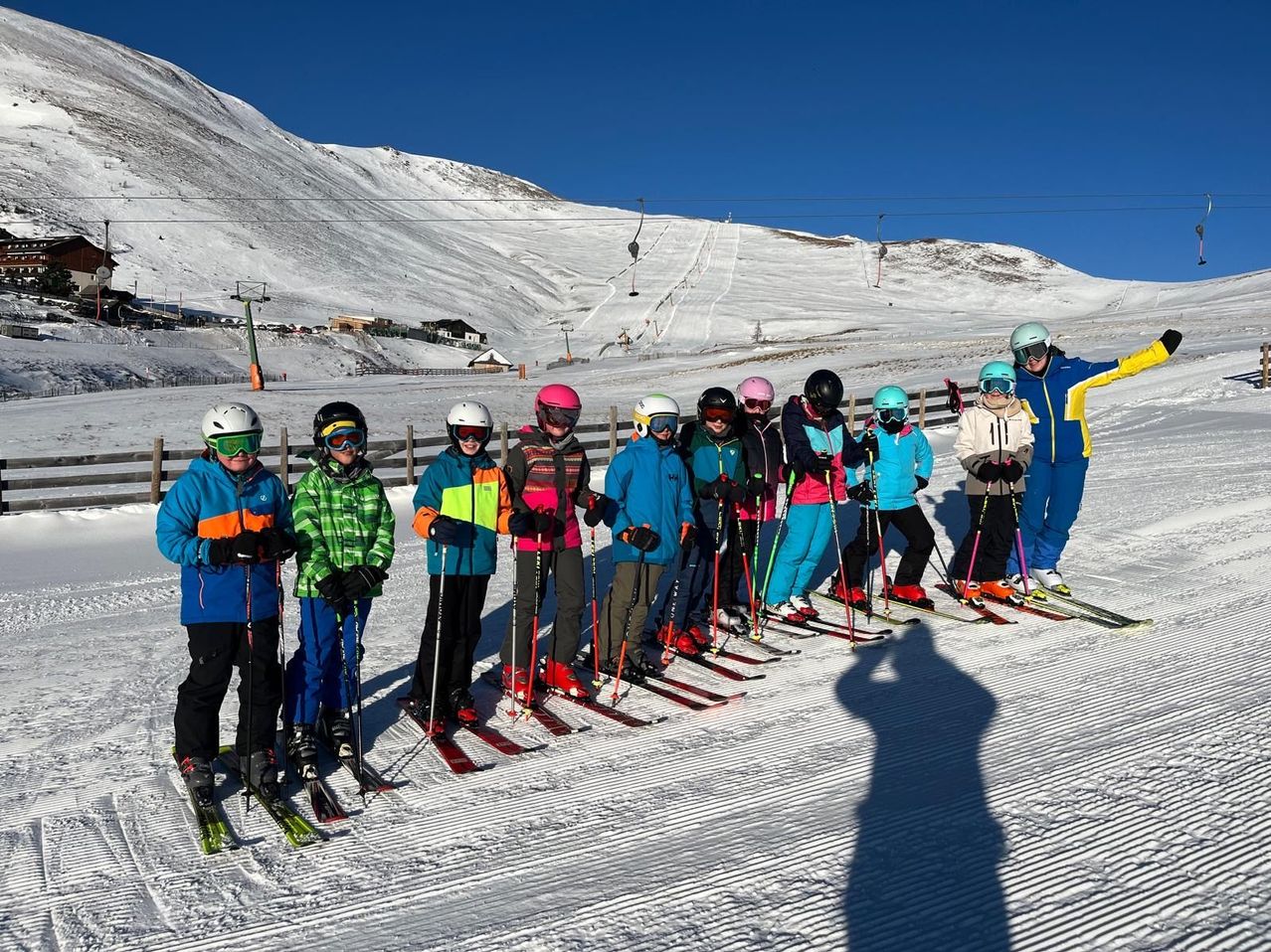 Eine Gruppe von Skifahrern posiert für ein Foto auf einem verschneiten Berg, mit klarem blauem Himmel oben und Skiliften im Hintergrund.
