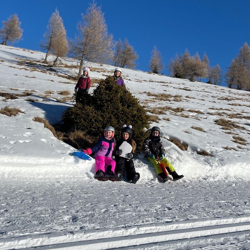 Fünf Kinder, alle in warme Kleidung gehüllt, posieren für ein Foto auf einem verschneiten Hügel. Drei sitzen im Schnee, eines steht hinter einem Busch und eines hält eine Schaufel.