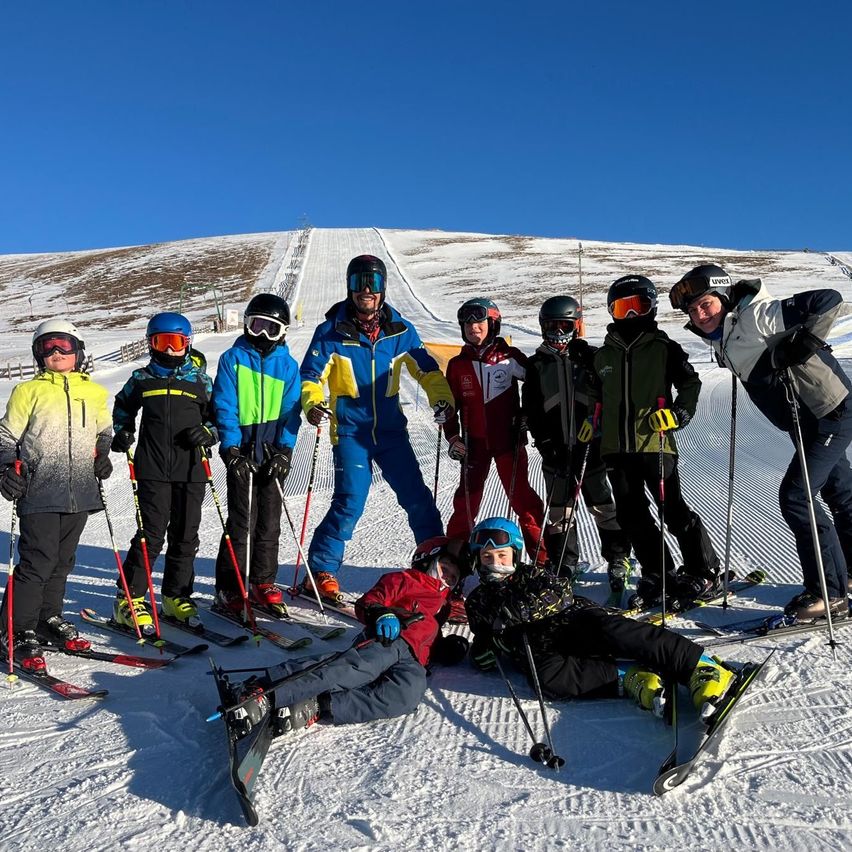Eine Gruppe von Skifahrern, einige stehend und andere sitzend, posiert auf einem verschneiten Hang bei blauem Himmel. Eine Person liegt im Schnee, und sie tragen alle Skiausrüstung.