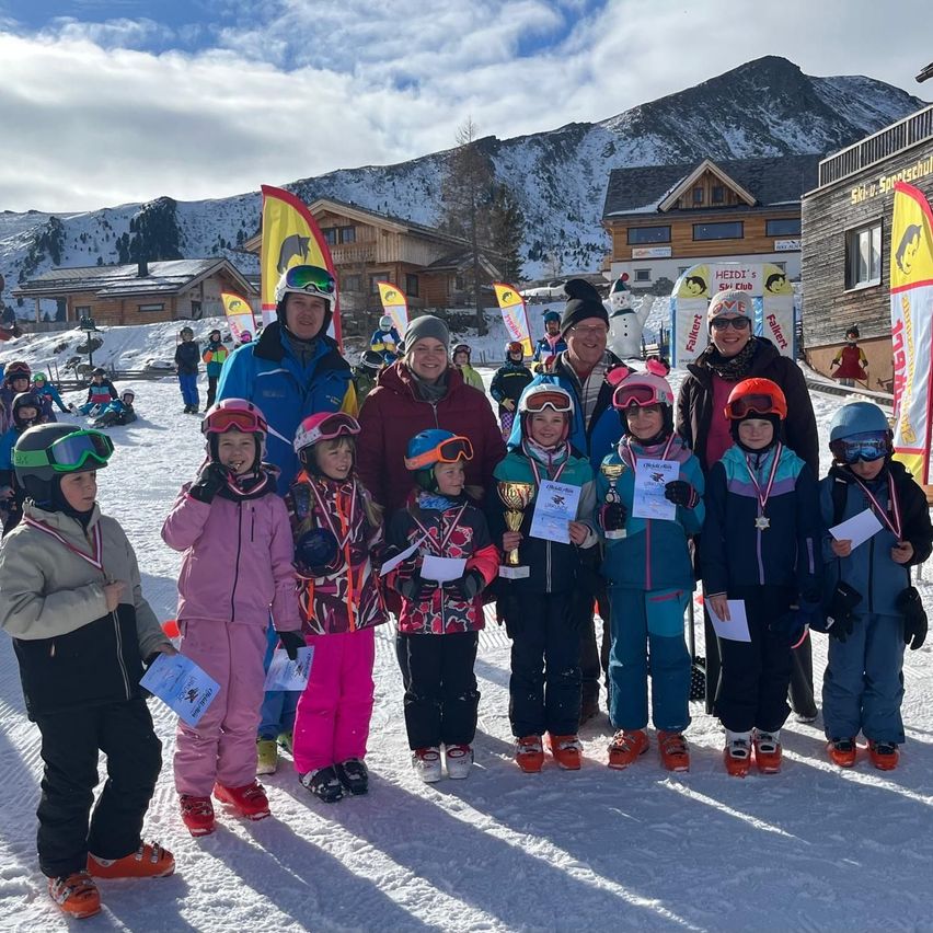 Eine Gruppe von Kindern und Erwachsenen steht auf einem verschneiten Feld. Sie halten Trophäen und tragen Winterkleidung. Hinter ihnen stehen einige Häuser mit Fahnen.