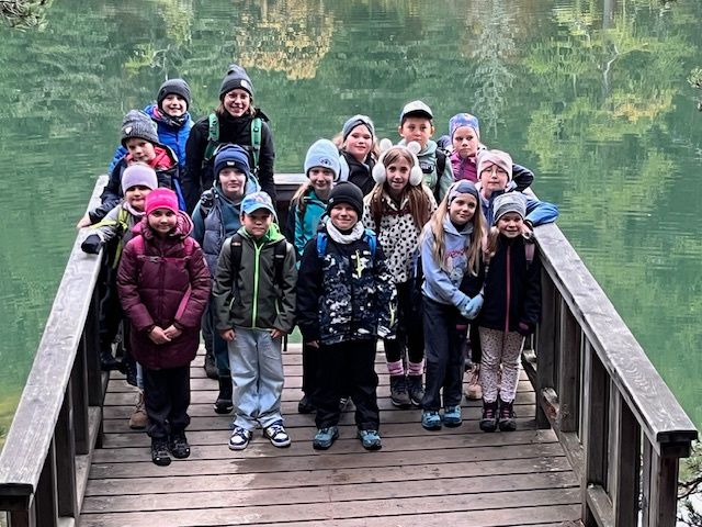 Eine Gruppe von Kindern in Winterkleidung steht auf einem Holzsteg am See und lächelt für ein Foto.
