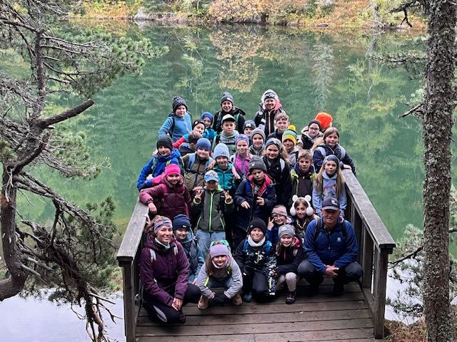 Eine Gruppe von Menschen, wahrscheinlich auf einer Wanderung, posiert für ein Foto auf einer Holzbrücke über einem See mit grünem Wasser und Bäumen.