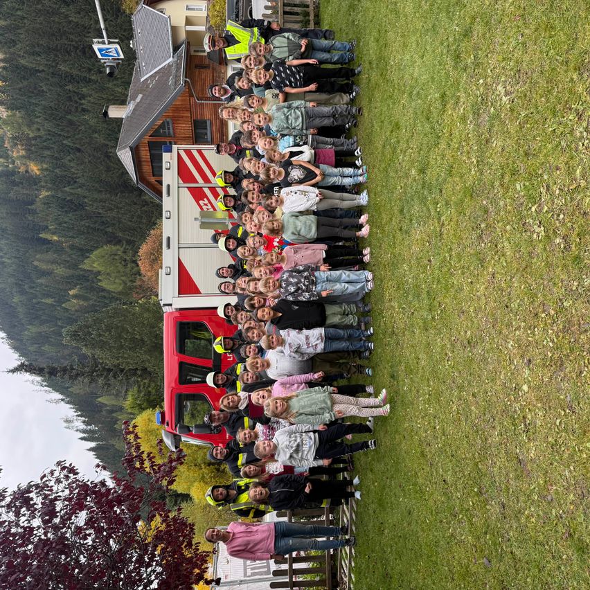 Eine große Gruppe von Kindern steht auf einem Rasenfeld, mit einem Feuerwehrauto dahinter. Sie sind in verschiedenen Outfits gekleidet und einige tragen reflektierende Westen. Im Hintergrund befindet sich ein Gebäude und Bäume.
