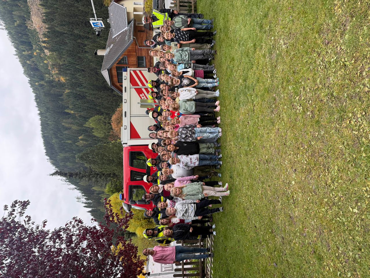 Eine große Gruppe von Kindern steht auf einem Rasenfeld, mit einem Feuerwehrauto dahinter. Sie sind in verschiedenen Outfits gekleidet und einige tragen reflektierende Westen. Im Hintergrund befindet sich ein Gebäude und Bäume.