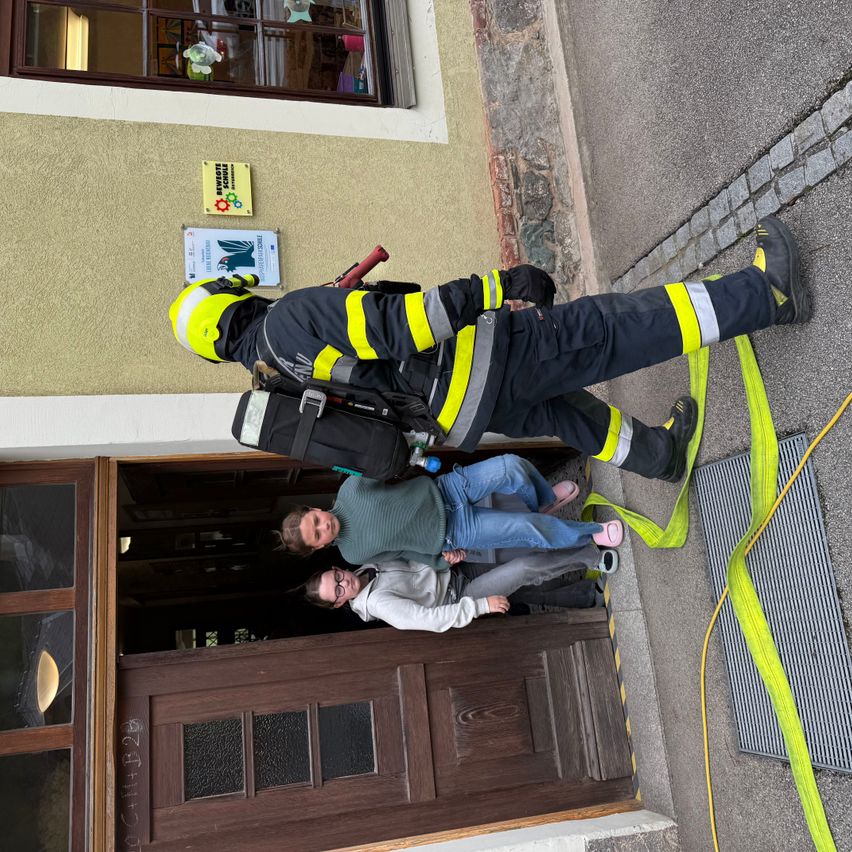 Ein Feuerwehrmann in voller Ausrüstung hilft zwei Frauen aus einem Gebäude. Die Frauen tragen Freizeitkleidung. Das Gebäude hat eine Holztür und Glasfenster.