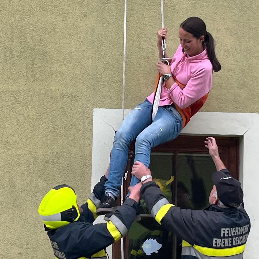 Eine Frau, die ein Seil hält, trägt ein rosa Shirt und Jeans und wird von zwei Feuerwehrleuten in gelben und schwarzen Uniformen hochgezogen.